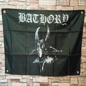 Può includere: Bandiera nera con la scritta "BATHORY" in lettere gotiche bianche sopra un'illustrazione bianca di una testa di capra. La bandiera ha occhielli in ottone in ogni angolo.