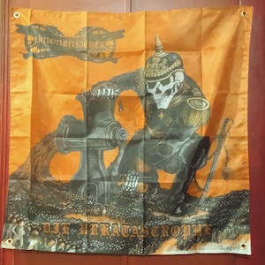 May include: Orange flag with a skeletal soldier in a Prussian helmet, operating a cannon. The flag has text at the top and bottom, and a cannon wheel graphic. The soldier is in a dark uniform with gold accents.