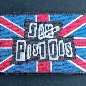May include: A rectangular embroidered patch featuring a Union Jack flag background in red, white, and blue. Centered on the patch is a black shape with the white text "SEX PISTOLS" written in a jagged, hand-drawn style font with a safety pin graphic.