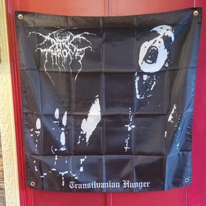 Puede incluir: Bandera negra con gráficos y texto en blanco. La bandera muestra el nombre de la banda "Darkthrone" en una fuente estilizada, una imagen de una cara gritando y el texto "Transilvanian Hunger". Tiene ojales metálicos en cada esquina.
