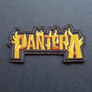 Peut inclure: Un écusson en tissu brodé affichant le mot PANTERA en lettres jaunes et orange en forme de flammes. Le texte est souligné par un fil noir, créant une forme distincte qui suit les contours des lettres sur un fond sombre et uni.