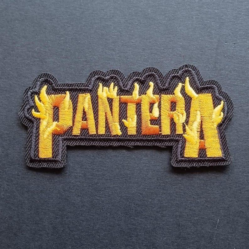 May include: An embroidered fabric patch featuring the word PANTERA in bold, yellow and orange flame-shaped lettering. The text is outlined in black thread, creating a distinct shape that follows the contours of the letters against a dark background.