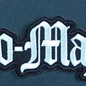Może przedstawiać: Haftowana naszywka z tkaniny z napisem Cro-Mags wykonanym stylizowaną białą czcionką gotycką z grubą czarną obwódką. Litery są wyszyte białą nicią na czarnym tle, co tworzy projekt o wysokim kontraście.