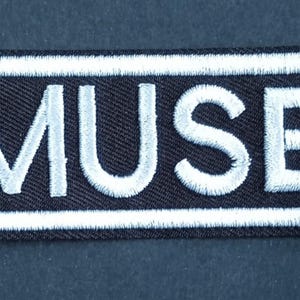 Può includere: Una toppa rettangolare ricamata con sfondo nero e la parola MUSE cucita in lettere maiuscole bianche. Il design include una linea di bordo orizzontale bianca che corre lungo i bordi superiore e inferiore della toppa.