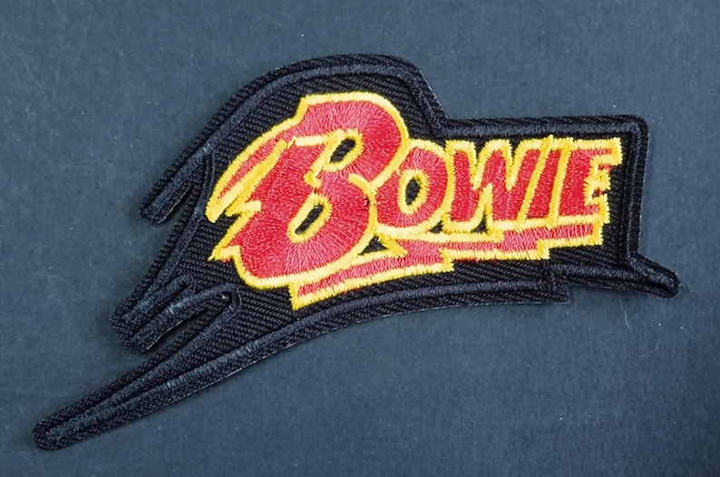 Peut inclure: Un &eacute;cusson brod&eacute; pr&eacute;sentant le mot Bowie en lettres rouges avec un contour jaune. Le texte est plac&eacute; sur un fond noir avec une forme stylis&eacute;e et dentel&eacute;e qui s'&eacute;tend vers la gauche, cr&eacute;ant un &eacute;l&eacute;ment de design angulaire et pointu.