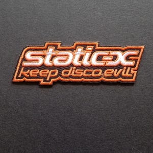 Peut inclure: Un écusson brodé présentant le texte "static-x" en blanc avec une bordure orange, placé au-dessus du texte "keep disco evil" en orange. Le motif a un fond noir et est souligné par une épaisse bordure brodée de couleur orange.