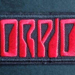 May include: A rectangular embroidered patch featuring the word SCORPIONS in bold, red, stylized capital letters. The lettering is set against a black background with a thick black stitched border surrounding the entire rectangular design.