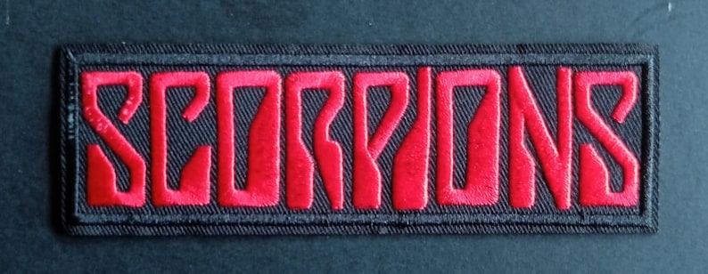 May include: A rectangular embroidered patch featuring the word SCORPIONS in bold, red, stylised capital letters. The lettering is set against a black background with a thick black stitched border surrounding the entire rectangular design.