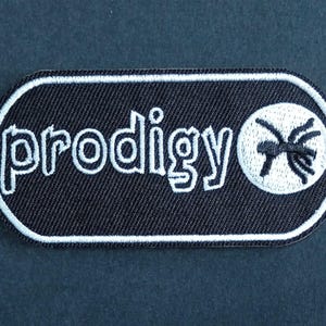 May include: A black rectangular embroidered patch with rounded corners and a white border. The patch features the white text "prodigy" in a stylized font followed by a white circular icon containing a black stylized ant graphic design.