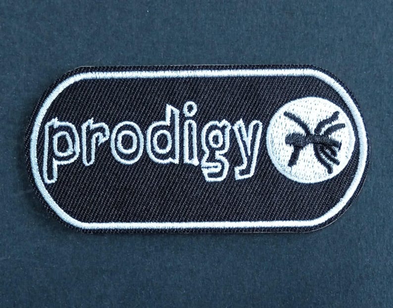 May include: A black rectangular embroidered patch with rounded corners and a white border. The patch features the white text "prodigy" in a stylized font followed by a white circular icon containing a black stylized ant graphic design.
