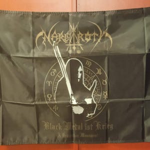 Puede incluir: Bandera negra con el nombre de la banda "Nargaroth" en letras doradas. Un gráfico central muestra una figura con una espada dentro de un círculo de símbolos astrológicos. El texto "Black Metal ist Krieg" está debajo.