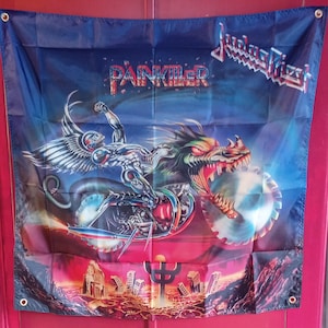 Puede incluir: Un estandarte de pared cuadrado de tela con la portada del álbum Painkiller de Judas Priest. Una figura cyborg metálica con alas monta una motocicleta con forma de dragón y ruedas de sierra circular sobre una ciudad en ruinas.