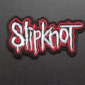 Peut inclure: Un écusson brodé présentant le mot Slipknot en lettres blanches stylisées avec un contour rouge. Le motif est placé sur un fond noir avec une bordure noire épaisse qui suit le contour irrégulier et dentelé du texte.