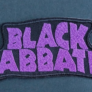May include: A rectangular embroidered patch with a black background and purple text that reads Black Sabbath. The patch features a thick black border and a textured surface created by the dense purple thread used for the lettering.