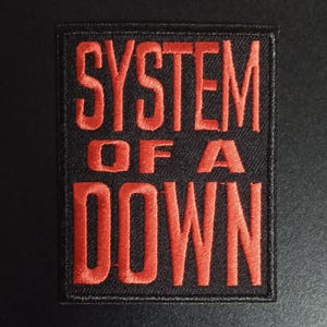 Peut inclure: Un écusson rectangulaire brodé présentant le texte SYSTEM OF A DOWN en lettres capitales rouges audacieuses sur un fond noir uni. Le lettrage est disposé sur trois lignes avec une bordure noire épaisse entourant tout le motif rectangulaire.