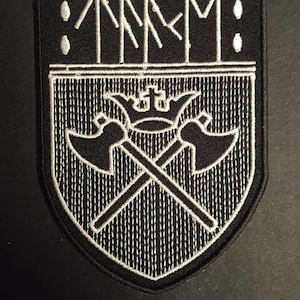 Peut inclure: Écusson brodé noir et blanc avec un motif de bouclier. L'écusson présente les lettres "TMM" au-dessus d'un motif de haches croisées avec une couronne. L'écusson est bordé de coutures blanches.