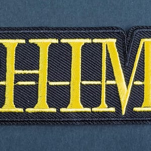 Könnte beinhalten: Ein rechteckiger gestickter Aufnäher mit dem Text HIM in fetten gelben Großbuchstaben. Die Buchstaben sind auf einen schwarzen Stoffhintergrund mit passendem schwarzem Rand gestickt. Das Design enthält eine horizontale Linie durch die Buchstaben.