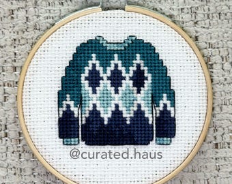 Peut inclure: Une broderie au point de croix dans un tambour en bois rond représentant un pull bleu sarcelle et bleu marine avec un motif losange sur une toile Aida blanche. Le texte @curated.haus est brodé en fil gris foncé sous le motif.