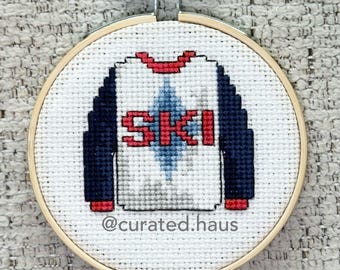 Peut inclure: Une broderie au point de croix dans un tambour en bois rond représentant un haut blanc à manches longues bleu marine, un col rouge et le mot SKI en lettres rouges sur un losange bleu clair. Le texte @curated.haus est en dessous.