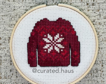 Peut inclure: Un tambour à broder circulaire en bois contenant une toile Aida blanche avec un pull rouge foncé brodé au point de croix, orné d'un motif de flocon de neige blanc au centre. Le texte @curated.haus est brodé en noir sous le pull.