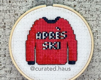 Peut inclure: Un tambour à broder circulaire en bois contenant une toile Aida blanche avec un motif au point de croix représentant un pull rouge à bordures bleu foncé. Le pull porte l'inscription "APRÈS SKI" en lettres blanches. "@curated.haus" est inscrit.