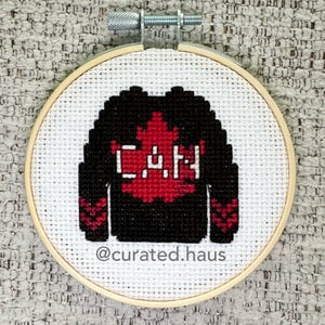 Könnte beinhalten: Ein Kreuzstich-Stickrahmen mit einem schwarzen Sweater-Design, einem roten Ahornblatt und den Buchstaben "CAN" in Weiß. Das Design befindet sich auf weißem Stoff, mit "@curated.haus" unten. Der Rahmen wird von einer Metallschraube gehalten.