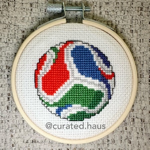 May include: A circular wooden embroidery hoop containing white Aida fabric with a cross-stitch design of a soccer ball featuring red, blue, and green panels outlined in white. The text @curated.haus is stitched in gray at the bottom center.
