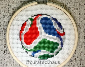 Peut inclure: Un tambour à broder circulaire en bois contenant une toile Aida blanche avec un motif au point de croix représentant un ballon stylisé aux segments rouges, bleus et verts bordés de gris. Le texte @curated.haus est brodé en gris en bas.