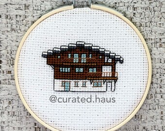 Peut inclure: Un tambour à broder circulaire en bois contenant une toile Aida blanche avec un motif au point de croix représentant un chalet brun et blanc à plusieurs étages. Le texte @curated.haus est brodé en fil gris foncé sous le bâtiment.