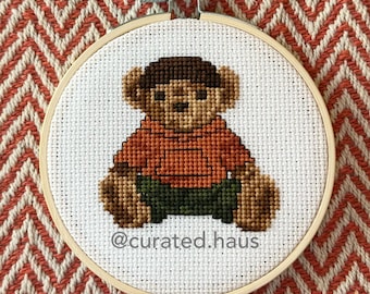 Peut inclure: Un tambour à broder circulaire en bois contenant un motif au point de croix représentant un ours en peluche marron portant un sweat à capuche orange et un pantalon vert foncé. Le texte @curated.haus est brodé en noir sous l'ours.