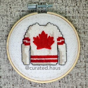 Peut inclure: Un cercle de broderie au point de croix représentant un maillot de hockey blanc avec une feuille d'érable et des rayures rouges. Le maillot est posé sur un fond de tissu blanc, encadré par un cercle en bois. Le texte "@curated.haus" est visible.