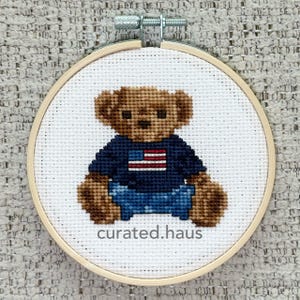 May include: A circular wooden embroidery hoop containing a cross-stitch design of a brown teddy bear wearing a navy blue shirt with a red and white striped flag graphic. The text curated.haus is stitched in grey below the bear on white fabric.