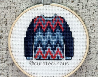 Peut inclure: Une broderie au point de croix représentant un pull avec un motif à chevrons rouge, bleu foncé et bleu clair, présentée dans un tambour en bois rond. Le tissu blanc porte le texte @curated.haus brodé en petites lettres noires.