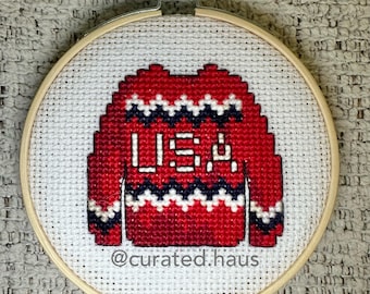 Peut inclure: Une broderie au point de croix dans un tambour en bois circulaire représentant un pull rouge avec des motifs en zigzag blancs et bleu marine. Le pull affiche le texte blanc USA au centre. Le texte @curated.haus est inscrit en dessous.