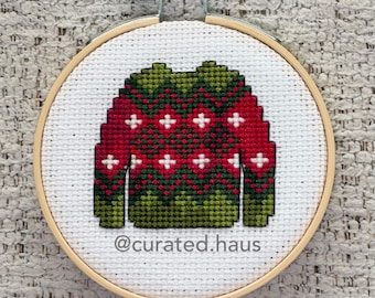 Peut inclure: Une broderie au point de croix dans un tambour en bois circulaire représentant un pull avec un motif géométrique rouge et vert et de petites croix blanches. Le tissu blanc affiche le texte @curated.haus en gris sous le pull.