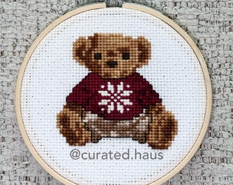Peut inclure: Une broderie au point de croix représentant un ours en peluche marron portant un pull rouge foncé avec un motif de flocon de neige blanc, encadrée dans un tambour en bois. Le texte @curated.haus est brodé en gris foncé sous l'ours.