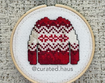Peut inclure: Une broderie au point de croix dans un tambour en bois rond représentant un pull rouge et blanc avec un motif géométrique. Le motif est centré sur une toile Aida blanche. Le texte @curated.haus est brodé en gris foncé en bas.