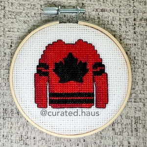 Op de afbeelding: Een borduurring met kruissteken die een rood hockey-shirt met een zwart esdoornblad en strepen voorstelt. De tekst "@curated:haus" is zichtbaar. De borduurring is een populair ambachtelijk item.