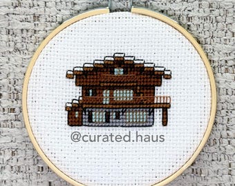Peut inclure: Une broderie au point de croix dans un cercle en bois représentant un chalet marron avec de la neige blanche sur le toit et des fenêtres bleues. Le tissu blanc affiche le texte @curated.haus en lettres grises sous le motif.