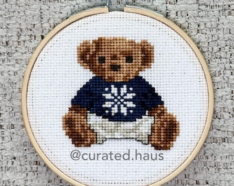 Peut inclure: Un tambour à broder circulaire en bois contenant un motif au point de croix représentant un ours en peluche marron portant un pull bleu foncé avec un motif de flocon de neige blanc et un pantalon blanc. Le texte @curated.haus est brodé en noir.