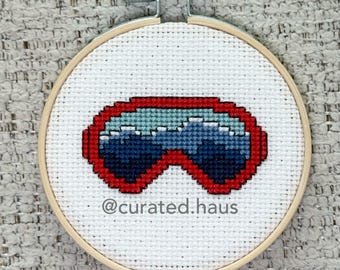 Peut inclure: Une broderie au point de croix dans un tambour en bois circulaire représentant une paire de lunettes à monture rouge avec des verres bleus. Le tissu Aida blanc affiche le texte @curated.haus en petites lettres noires sous les lunettes.