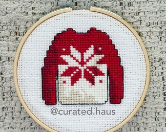 Peut inclure: Une broderie au point de croix dans un tambour en bois rond représentant un pull rouge avec un motif de flocon de neige blanc sur une toile Aida blanche. Le texte @curated.haus est brodé en fil noir au bas du motif en tissu.