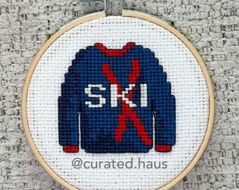 Peut inclure: Un tambour à broder circulaire en bois contenant un tissu Aida blanc avec un pull bleu foncé brodé au point de croix, orné d'un X rouge et du texte blanc SKI. Le texte @curated.haus est brodé en gris au bas du motif.
