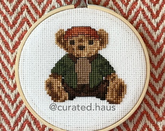 Peut inclure: Une broderie au point de croix dans un tambour en bois circulaire représentant un ours en peluche marron portant une veste vert foncé et une casquette brun rougeâtre. L'ours est centré sur un tissu blanc avec le texte @curated.haus en gris.
