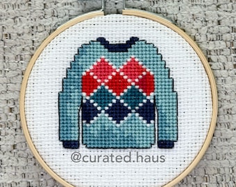 Peut inclure: Un tambour à broder circulaire en bois contenant du tissu Aida blanc avec un pull au point de croix présentant un motif à losanges argyle rouge, bleu sarcelle et bleu marine. Le texte @curated.haus est brodé en fil bleu foncé sous le pull.