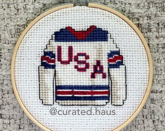 Peut inclure: Broderie au point de croix d'un maillot de hockey USA dans un cercle en bois. Le maillot est blanc avec des accents rouges et bleus, et les lettres "USA" sont brodées en rouge. La broderie est posée sur un fond en tissu neutre.