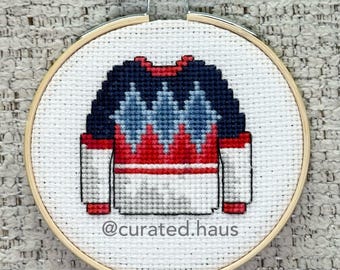 Peut inclure: Une broderie au point de croix d'un pull dans un tambour en bois. Le pull présente une partie supérieure bleu foncé avec des motifs en losanges bleu clair, une bande centrale rouge et une partie inférieure blanche. Le texte @curated.haus est inscrit.
