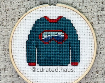 Peut inclure: Une broderie au point de croix dans un tambour en bois circulaire représentant un pull bleu sarcelle foncé avec des bordures rouges et un motif central de lunettes bleues et blanches. Le tissu blanc porte le texte @curated.haus en noir.