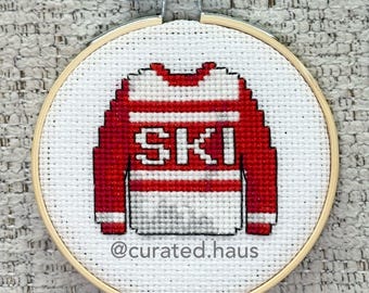 Peut inclure: Un tambour à broder circulaire en bois contenant une toile Aida blanche avec un motif au point de croix représentant un pull rouge et blanc avec le texte SKI au centre. Le texte @curated.haus est brodé en gris au bas de la toile.