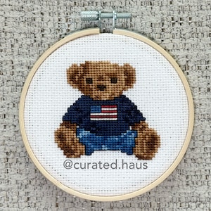 May include: A cross-stitch embroidery in a circular wooden hoop featuring a brown teddy bear wearing a dark blue shirt with a red and white American flag design. The text @curated.haus is stitched in dark gray at the bottom of the white fabric.